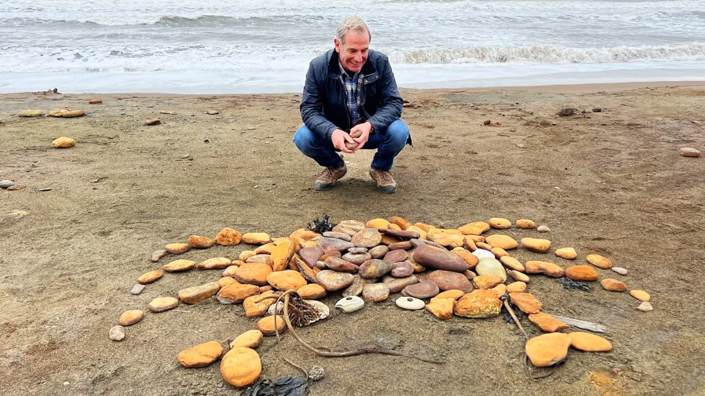 Robson Green on a beach looking down at a crab design made out of rocks