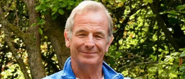 Robson Green in a blue shirt with trees behind him