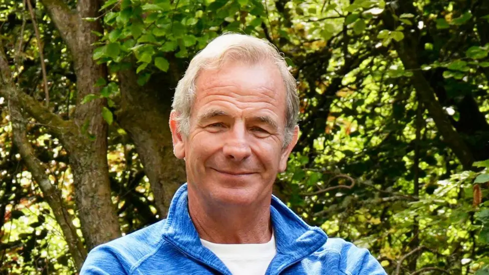 Robson Green in a blue shirt with trees behind him